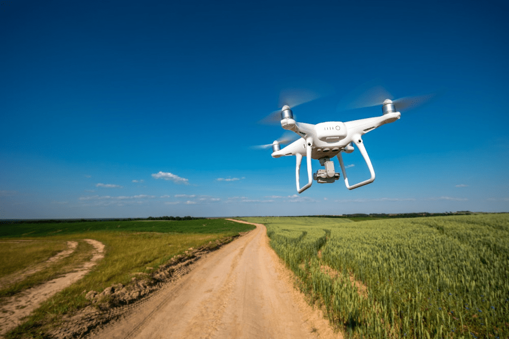 White drone flying over a dirt road and green fields, representing GPS tracking for drones and aerial fleet management.