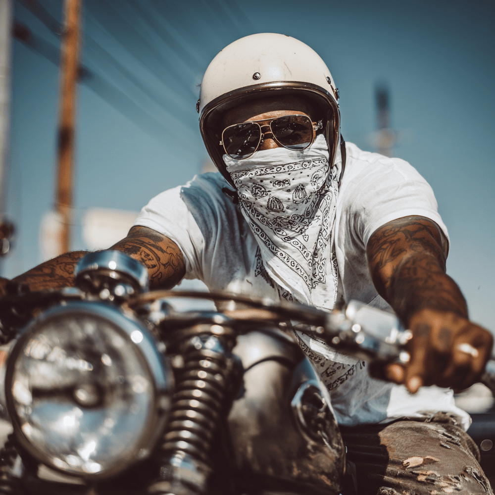 Person riding a motorcycle wearing a helmet, sunglasses, and bandana with a clear blue sky in the background.
