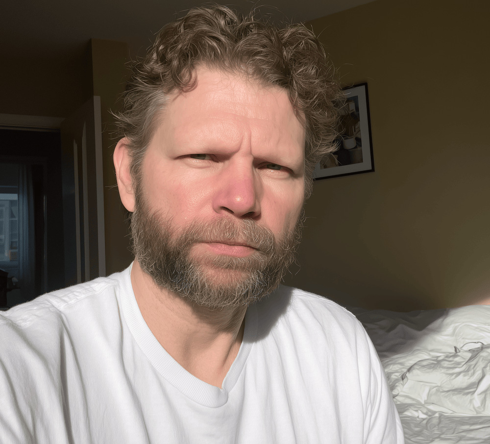 Man with a beard wearing a white shirt in a room with a bed and framed picture on the wall.
