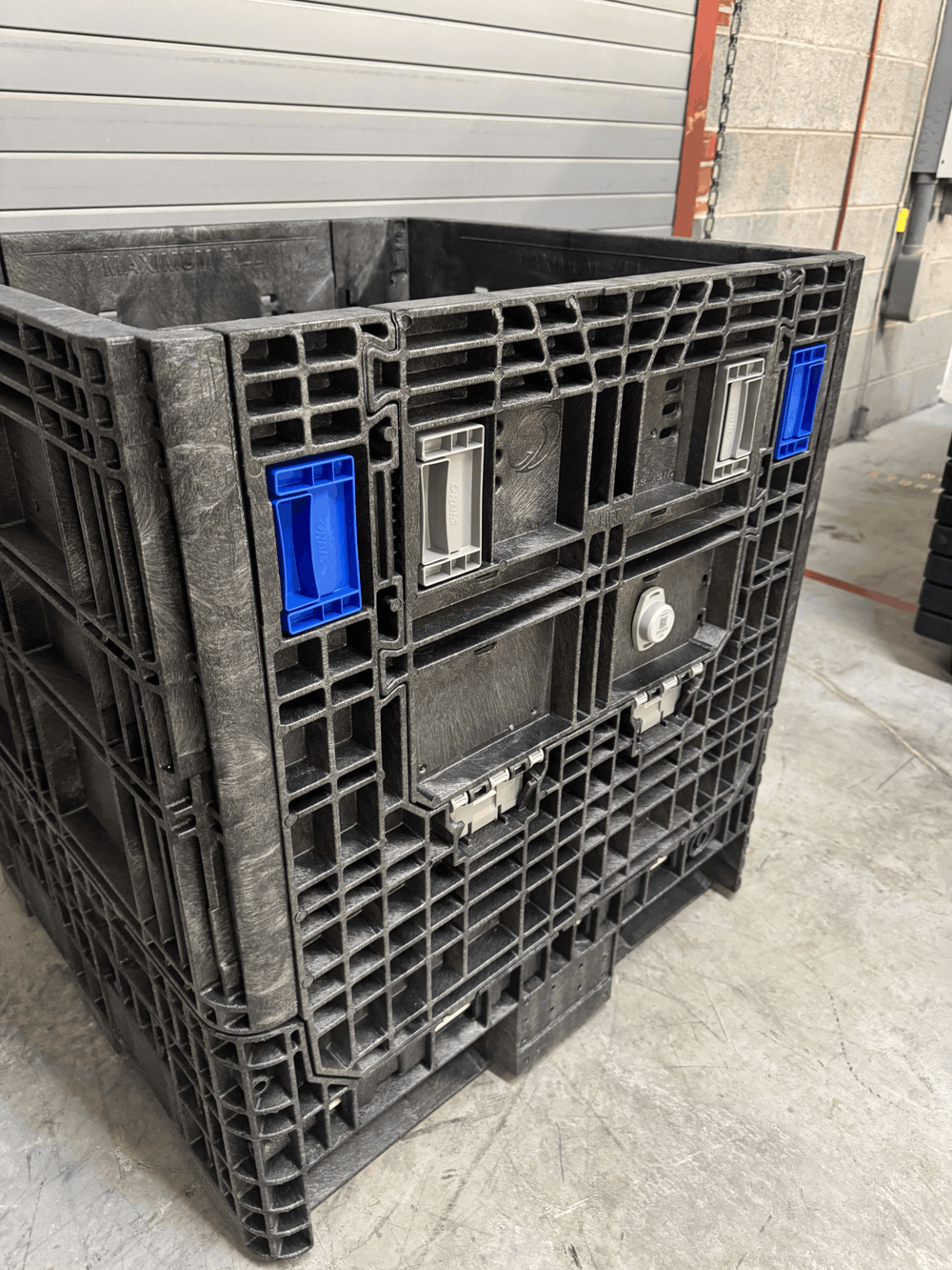 Tracking device mounted on a large black plastic pallet crate with integrated latches and blue plastic handle inserts, photographed in a warehouse setting.