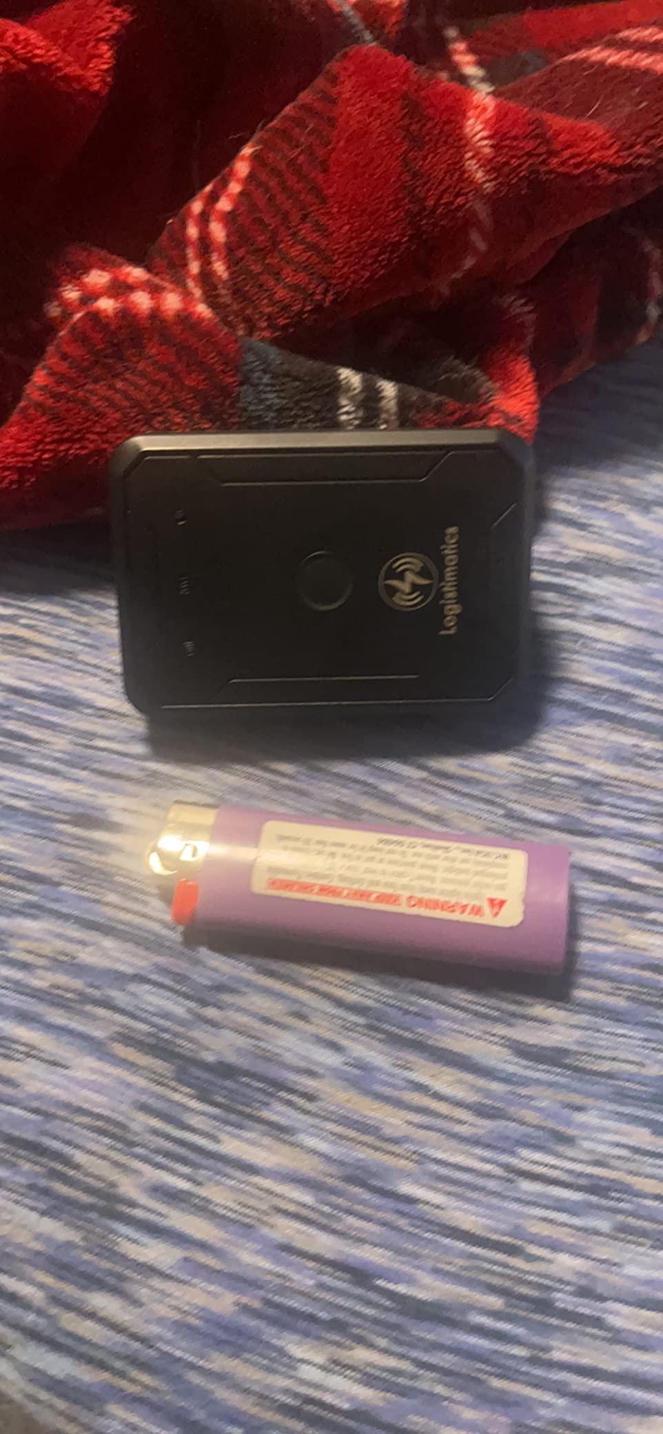 Small black GPS device and purple Bic Lighter on a bed with a red blanket in the background. Showing size comparison to lighter. 