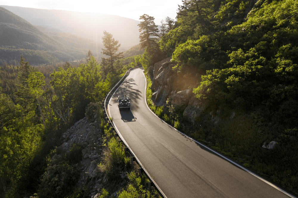 Convertible car driving along a winding mountain road surrounded by lush green forest and rocky cliffs, showcasing scenic travel and adventure.